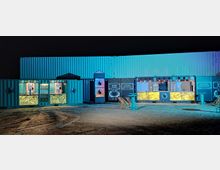 Eine nächtliche Szene mit beleuchteten Containern, die als Bar- und Loungebereich umfunktioniert wurden. Die Container verfügen über Tresenelemente mit gelblicher Beleuchtung, Getränkeauslagen und Sitzgelegenheiten auf einem sandigen Boden davor.