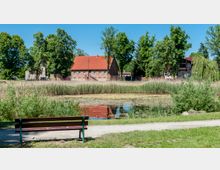 Parklandschaft mit Teich im Vordergrund, umgeben von Schilf und Büschen; im Hintergrund stehen mehrere Gebäude mit rotem Ziegeldach, darunter ein Fachwerkhaus, eingebettet in hohe Bäume. Eine Holzbank steht auf einem Kiesweg vor dem Teich.