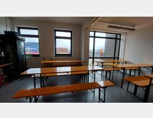 Ein einfacher Raum mit mehreren hölzernen Bierbänken und Tischen, hell beleuchtet durch Fenster an zwei Seiten. Draußen sind Schiffscontainer und ein Industriegelände zu sehen, und ein Kühlschrank steht an der linken Wand.