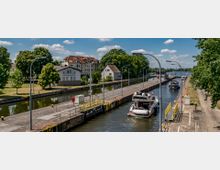 Schleusenanlage mit moderner Infrastruktur, durch die mehrere Boote fahren; im Hintergrund Wohnhäuser, Bäume und ein Fluss unter blauem Himmel mit wenigen Wolken.