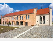 Backsteinbau mit roten Dachziegeln und großen Rundbogenfenstern, umgeben von einem gepflasterten Hof mit moderner Rampe und Fahrradständern; im Hintergrund ein blauer Himmel mit Wolken.