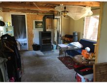 Ein einfacher Innenraum mit einem Holzofen als zentralem Element, einer blauen Couch, einem kleinen Tisch und Regalen. Sichtbare Holzbalken an der Decke und eine Tür im Hintergrund führen in einen Nebenraum.