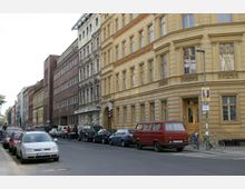 Straßenszene mit mehrstöckigen Altbauten auf der rechten Seite, teils mit gelber Fassade und verzierten Fenstern, daneben Backstein- und Putzgebäude. Auf der Straße parken mehrere Autos und ein roter Kleinbus, während Fahrräder an einem Straßenlaternenpfosten angelehnt sind.