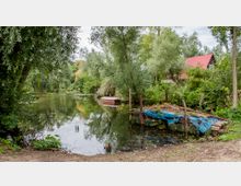 Ein ruhiger Flusslauf mit dichtem, grünem Uferbewuchs, einem kleinen roten Boot am Wasser und einer improvisierten Brücke aus Holz und blauen Planen. Im Hintergrund steht ein Haus mit einem roten Satteldach zwischen den Bäumen.
