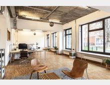 Ein modernes Büro mit einem offenen Grundriss, Holzboden und großen Fenstern, die viel Tageslicht hereinlassen. Der Raum ist mit Schreibtischen, Stühlen und minimalistischen Möbeln ausgestattet, und die Decke zeigt freiliegende Rohre und Beleuchtung.