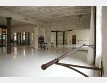 Ein Tanzstudio in einem industriellen Gebäude mit sichtbaren Backsteinwänden und einer hohen Decke. Der Raum verfügt über Spiegelwände, Balletstangen, einen glänzenden weißen Boden und große Fenster mit Blick auf den Außenbereich.