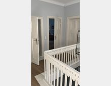 Ein heller Innenbereich eines Hauses mit einem weißen Holzgeländer im Vordergrund, das einen Treppenaufgang umgibt. Im Hintergrund führen drei Türen zu verschiedenen Räumen, von denen eine leicht geöffnet ist und einen Blick auf ein blau gestrichenes Zimmer mit einem Sofa erlaubt.