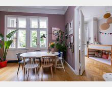 Essbereich in einem Wohnzimmer mit einem runden weißen Esstisch und unterschiedlichen Stühlen vor einer großen Fensterfront. Eine lila Wand ist mit Bildern dekoriert, Pflanzen stehen auf dem Boden, und rechts führt eine Tür in ein Kinderzimmer mit einem Spielbett und bunten Dekorationen.