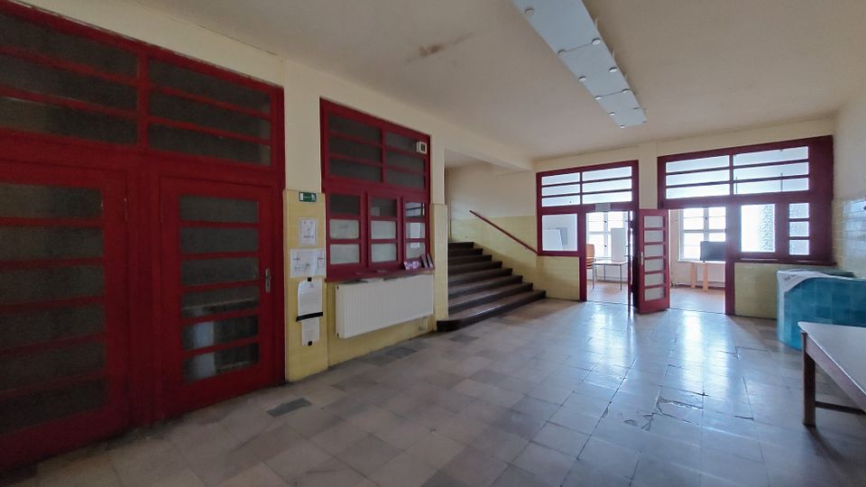 Flur eines Gebäudes mit roten Tür- und Fensterrahmen, gefliesten gelben Wänden und einer Treppe, die nach oben führt. Im Hintergrund sind Räume mit großen Fenstern und Möblierung erkennbar.