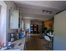 Ein offener Raum, der eine kleine Küche mit Arbeitsfläche, Kaffeemaschinen und Hängeschränken links zeigt, sowie eine Sitzecke mit hohen Hockern rechts. Im Hintergrund ist eine Backsteinwand und professionelle Audio- oder Technikausrüstung zu sehen.