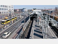 Straßenansicht mit einer erhöhten U-Bahn-Station auf der rechten Seite, darunter Bahngleise, Laternen und ein Bahnsteigdach. Links befindet sich eine Straße mit gelben Straßenbahnen, einem Taxi und Fußgängern; im Hintergrund sind mehrstöckige Gebäude und ein Baukran sichtbar.