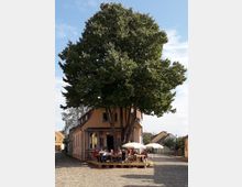 Ein kleiner Platz mit Kopfsteinpflaster, darauf ein Café mit einer Holzbühne unter einem großen Baum. Gäste sitzen an Tischen mit weißen Sonnenschirmen vor einem gelb-orangefarbenen Gebäude.