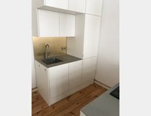 Kleine, minimalistische Einbauküche mit weißen Schränken, grauer Arbeitsplatte, Edelstahlspüle und Holzfußboden. Die Rückwand der Küche ist mit einer goldfarbenen Oberfläche versehen.