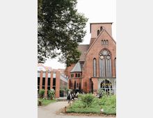 Backstein-Kirche mit großen gotischen Fenstern, flankiert von einem modernen Anbau mit Glas- und Backsteinelementen; im Vordergrund ein Grünbereich mit Blumen, Spazierwegen und sitzenden Personen.