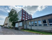 Mehrstöckiges Industriegebäude mit roten und grauen Fassaden sowie großen Fenstern, flankiert von einem niedrigeren, flachen Anbau mit Gitterfenstern. Im Vordergrund ein gepflasterter Hof, links ein hoher Baum, im Hintergrund blauer Himmel mit wenigen Wolken.