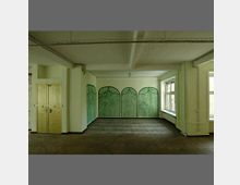 Ein leerer Raum mit Holzparkettboden, hellgrünen Wänden und einer dekorativen Wandgestaltung mit gemalten, gewölbten Fensterformen. Rechts sind zwei große Fenster zu sehen, links eine doppelflügelige gelbe Tür.