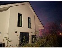 Zweigeschossiges modernes Wohnhaus mit weißer Fassade, großen Fenstern und einem geneigten Dach. Im Vordergrund stehen Pflanzen, während der Hintergrund einen Nachthimmel und Bäume zeigt.