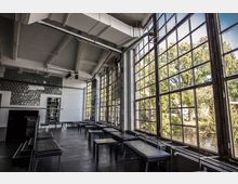 Großzügiger, lichtdurchfluteter Raum mit industriellem Design, gekennzeichnet durch hohe Decken mit sichtbaren Rohren, große, gitterartige Fenster und dunkle Sitzbänke sowie Tische. Durch die Fenster ist eine Grünfläche mit Bäumen und einem Gewässer sichtbar.