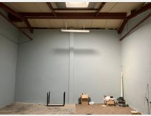 Empty warehouse-style interior with tall gray walls, exposed red steel ceiling beams, and a hanging fluorescent light fixture. Concrete floor area meets a section of wood-pattern flooring, with a few boxes and coiled cables stacked along one side.