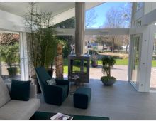 Ein moderner Wintergarten mit großen Glasfenstern und Blick auf eine grüne Außenanlage. Im Raum stehen Polstermöbel, ein Kaminofen, Pflanzen in Töpfen und ein Stapel Kaminholz.