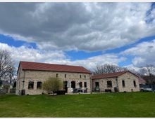 Steinhaus im Landhausstil mit roten Ziegeldächern, umgeben von einer großen Rasenfläche und kahlen Bäumen im Hintergrund. Der Himmel ist teils bewölkt mit blauen Abschnitten.