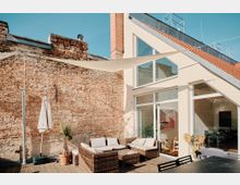 Sonnige Dachterrasse mit einer Sitzgruppe aus Rattanmöbeln, einem Sonnenschirm und einer Topfpflanze vor einer Backsteinwand. Im Hintergrund ein modernes Gebäude mit großen Glasfenstern und einem roten Ziegeldach.