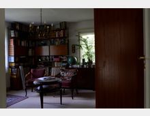 Wohnzimmer mit Bücherregalen an der Wand, gefüllt mit Büchern und Aktenordnern. Im Vordergrund stehen zwei gepolsterte Sessel, ein runder Beistelltisch und im Hintergrund ein Fenster mit einer Zimmerpflanze und einem Globus auf einer Kommode.