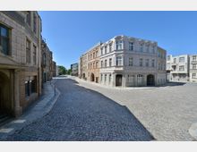 Kopfsteinpflasterstraße in einer historischen Altstadt mit mehrstöckigen, hellen Gebäuden im Stil des 19. Jahrhunderts; im Hintergrund Bäume und moderneres Gebäude sichtbar, unter klarem, blauem Himmel.