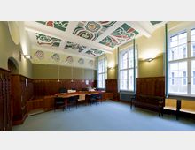 Raum mit hohen Decken und großen Fenstern, geschmückt mit dekorativ bemalter Stuckverzierung. Holzwände mit gotischen Ornamenten umgeben einen Besprechungstisch mit Stühlen, während die Wandfarbe und Lichtgestaltung eine helle, klassische Atmosphäre erzeugen.