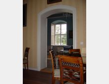 Ein Innenraum mit Holzboden und verzierten Holzstühlen, im Vordergrund gelbe Tapete. Ein offener Rundbogen führt zu einem angrenzenden Raum mit einem großen Fenster und Blick ins Freie.