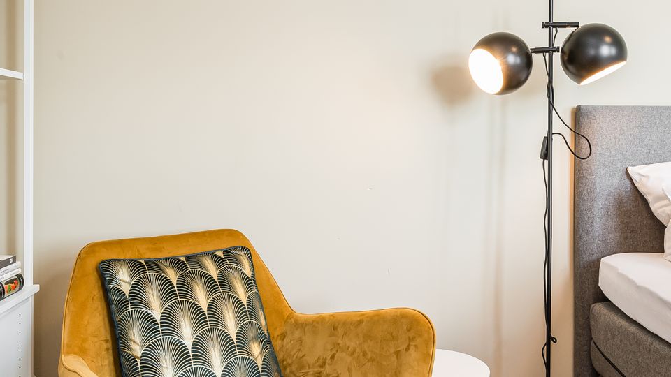 Gemütliche Zimmerecke mit einem senfgelben Sessel und gemustertem Kissen auf Parkettboden, daneben ein kleiner runder Beistelltisch und eine schwarze Stehlampe mit zwei kugelförmigen Leuchten. Am Rand sind ein weißes Regal und ein Teil eines Betts mit grauem Kopfteil zu sehen.