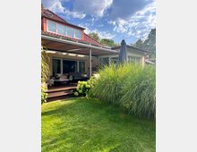 Rückseite eines Einfamilienhauses mit rotem Ziegeldach und großer, überdachter Holzterrasse mit Sitzbereich und Glasschiebetüren. Im Vordergrund liegt ein gepflegter Rasen mit hohen Ziergräsern, einer Hortensie und einem geschlossenen Sonnenschirm unter blauem Himmel mit Wolken.