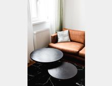 Wohnzimmer-Ecke mit einem braunen Ledersofa, einem weißen Kissen, zwei runden schwarzen Couchtischen und einem schwarzen Teppich mit weißem Muster; im Hintergrund ein Fenster mit weißen und grünen Vorhängen sowie ein Heizkörper.