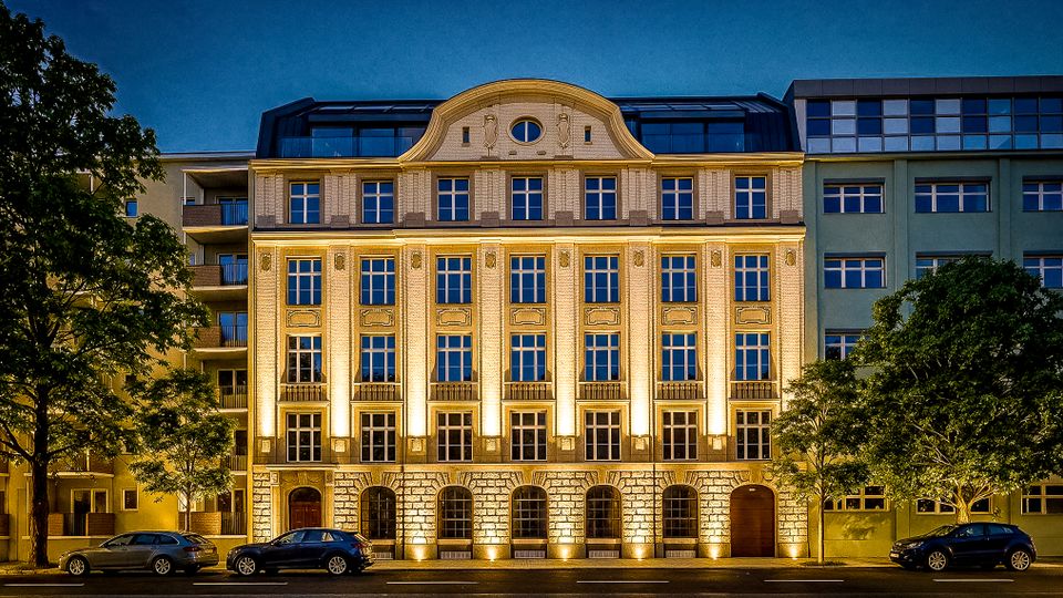 Beleuchtete historische Gebäudefassade in einer Stadtstraße bei Nacht, mit symmetrischen Fensterreihen, verziertem Mittelgiebel und einem rustizierten Sockel mit Rundbogenöffnungen. Vor dem Haus stehen Bäume und parkende Autos, rechts und links schließen moderne Nachbargebäude an.