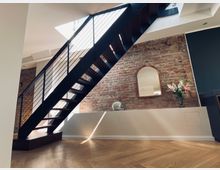 Modernes Innenraumdesign mit einer schwarzen Metalltreppe, die schräg nach oben verläuft, einer freiliegenden Ziegelwand im Hintergrund und einem dekorativen Spiegel sowie einem Blumenstrauß auf einer weißen Ablage. Der Boden besteht aus hellem Fischgrätparkett, und Tageslicht fällt durch ein Dachfenster ein.