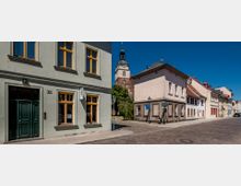 Straße in einer Altstadt mit historischen Gebäuden, darunter ein hellgrünes Haus mit gelben Fensterrahmen und ein beiges Haus. Im Hintergrund ist der Turm einer Backsteinkirche mit schwarzer Kuppel zu sehen, umgeben von Bäumen. Der Himmel ist klar und blau.