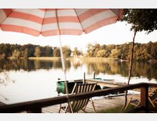 Ein Holzsteg an einem See mit Blick auf einen bewaldeten Uferbereich im Hintergrund. Im Vordergrund stehen ein gestreifter Liegestuhl und ein rot-weiß gestreifter Sonnenschirm, daneben ist ein kleines Ruderboot zu sehen.