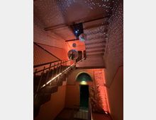 Treppe in einem Gebäude mit dekorativer Beleuchtung. Mehrere Discokugeln hängen von der Decke, und Lichtpunkte reflektieren an den Wänden und der Decke; ein beleuchteter Ausgang mit Rundbogenfenster befindet sich im Hintergrund.