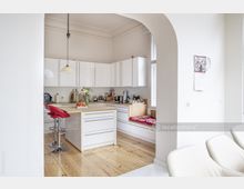 Helle, moderne Küche mit weißen Hochglanzschränken, einer Kücheninsel mit Holzarbeitsplatte und zwei roten Barhockern. Im Hintergrund befinden sich ein integrierter Sitzbereich mit Kissen und große Fenster, die für viel Tageslicht sorgen.