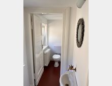 Kleines Badezimmer mit weiß gestrichenen Wänden, rotem Boden und einer weißen Toilette. Im Vordergrund sind ein Wandwaschbecken mit Messingarmatur und ein runder Wandspiegel sichtbar.