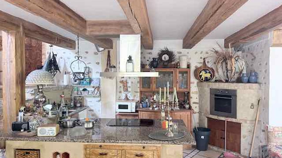 Rustikale Landhausküche mit freiliegenden Holzdeckenbalken, großer Kücheninsel aus Naturstein und Holz sowie vielen Küchenutensilien auf der Arbeitsfläche. Im Hintergrund sind ein Holzschrank, ein eingebauter Backofen und helle Fliesen mit gemustertem Rand zu sehen.