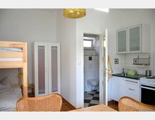 Kompakter Wohnraum mit Kochnische, Esstisch und Etagenbett. Im Hintergrund eine Tür, die zu einem kleinen Badezimmer mit gefliesten Wänden führt, sowie ein Kleiderschrank und Küchenmöbel mit Herd, Spüle und Hängeschränken.