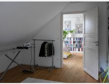 Ein Raum mit Holzfußboden und einer Dachschräge, ausgestattet mit einem Bügelbrett, einer Kleiderstange und einem offenen weißen Türrahmen. Hinter der Tür ist ein weiterer Raum mit Bücherregalen, Pflanzen und zwei Wandbildern zu sehen.