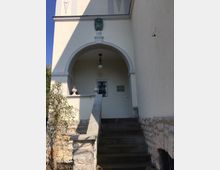 Eingang eines historischen Gebäudes mit einem Rundbogen, einer Steintreppe und einer kleinen Lampe unter dem Bogengewölbe. Die Fassade ist hell verputzt, und an der Wand befindet sich ein Schild sowie ein Löwenkopfrelief oberhalb des Bogens.