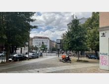 Straßenszene in einer Stadt mit gepflastertem Gehweg, mehreren Bäumen, parkenden Autos und Fahrrädern. Im Hintergrund stehen mehrstöckige Wohngebäude, und eine Uhr auf einer Werbetafel ist in der Mitte des Bildes sichtbar.