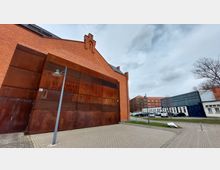 Backstein-Gebäude mit einem modernen, rostfarbenen Metallelement an der Fassade, nebendran eine gepflasterte Fläche und ein Parkplatz. Im Hintergrund sind weitere Gebäude im ähnlichen Stil sowie kahle Bäume zu sehen, unter einem grauen Himmel.