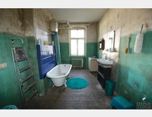 Ein einfaches Badezimmer mit abgenutzten Wänden in Grün- und Beigetönen. Es verfügt über eine freistehende Badewanne, ein Waschbecken, einen Spiegel, einen Heizkörper unter einem großen Fenster sowie blaue Fliesen an der Wand hinter der Badewanne. Auf dem Holzboden liegt ein türkisfarbener Teppich.