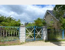 Ein Eingangstor aus blauem Metall mit weißen Elementen, eingerahmt von gemauerten Steinsäulen, führt zu einem Vorgarten mit dichtem Grün. Im Hintergrund ist ein Einfamilienhaus aus hellem Klinker mit dunklem Schrägdach sichtbar.