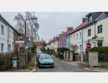 Straßenszene in einem Wohngebiet mit Reihenhäusern, die in verschiedenen pastellfarbenen Fassaden gestrichen sind. Die Straße ist schmal, mit parkenden Autos an einer Seite, niedrigen Zäunen und vereinzelten Bäumen entlang der Gehwege. Ein Straßenschild mit der Aufschrift „Bleicheroder Straße“ und ein Verkehrsschild sind im Vordergrund sichtbar.