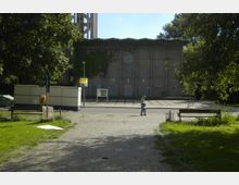 Außenansicht eines modernen Kirchengebäudes mit einer betonierten Fassade und einem runden Fenster, umgeben von Bäumen und Grünflächen. Im Vordergrund führt ein gepflasterter Weg mit einer Sitzbank zum Gebäude; eine Person geht in Richtung des Eingangs.